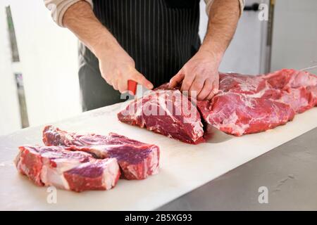 Butcher or cook slicing chuck steaks, cutting the big piece of fresh meat. Close-up view of male hands with a knife in the kitchen Stock Photo