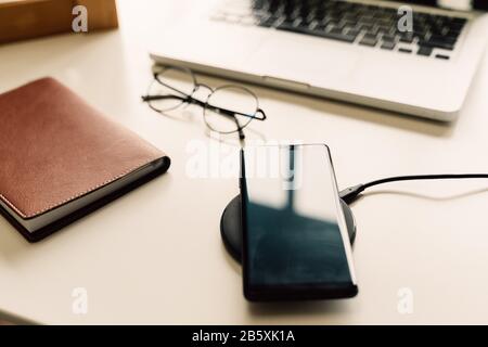 Smart phone being charged on wireless charger alongside with laptop, glasses, and notebook on table - Modern technology lifestyle concept Stock Photo