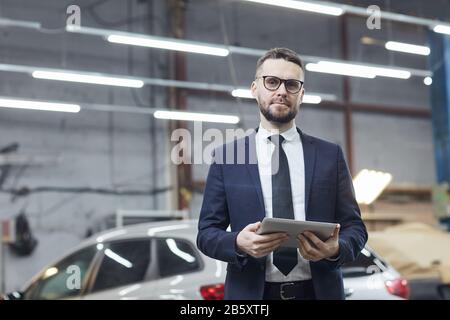Horizontal medium portrait of handsome Caucasian businessman standing in auto service center with tablet PC looking at camera Stock Photo