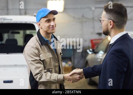 Horizontal medium portrait shot of professional auto mechanic greeting client shaking hands with him in car repair shop Stock Photo