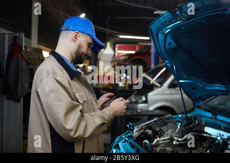 Cuacsian man with beard on face working in auto service workshop using tablet PC, horizontal side view shot Stock Photo