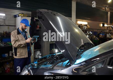 Horizontal medium long shot of modern auto mechanic using tablet PC while working in auto service workshop, copy space Stock Photo
