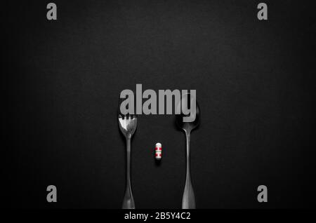 Fork and spoon with a capsule medicine on dark background. Minimalist flat lay black for health care concept. Stock Photo