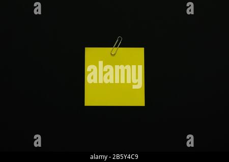 Yellow blank note paper with paper clip on dark background. Minimalist flat lay black concept. Stock Photo