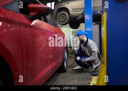 Horizontal side view shot of professional repairman sitting in front of car wheel using tire pressure gage Stock Photo