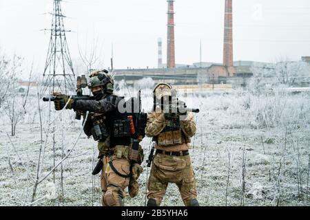 Men in camouflage cloth and black uniform with machineguns with factory on background. Soldiers with muchinegun aims aiming. Stock Photo