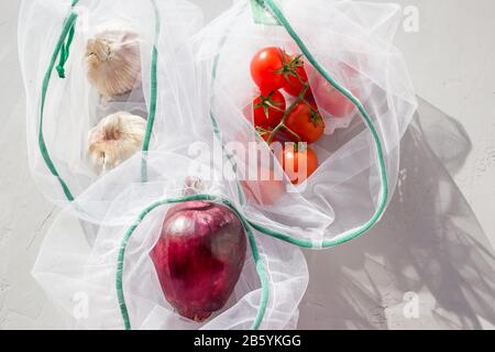 The reusable organza net bag for shopping with tomatoes, onion and garlic on grey background. Concept of no plastic, zero waste, reusable life. Stock Photo