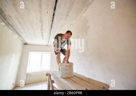 Male is making repairing in apartment by putting putty on the walls and standing on wooden stand against a window. Young man with a beard in a T-shirt and shorts smeared with paint Stock Photo