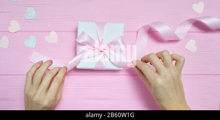 Valentine love concept of beautiful womans hands hold gift box craft paper and heart confetti on pink background. top view flat lay with place for Stock Photo