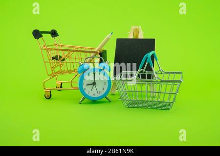 Shopping concept with shopping cart, basket and alarm clock, small chalk board over a green background Stock Photo