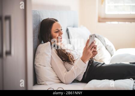 Happy businesswoman on business trip lying in hotel room and using smartphone. Stock Photo