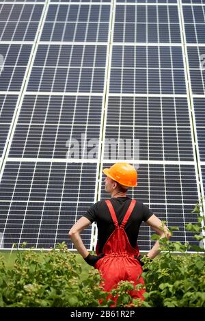 Back view of worker in orange uniform and helmet standing against the background of large plantation solar panels. Home construction of solar station. Vertical shot. Green ecological energy generation Stock Photo