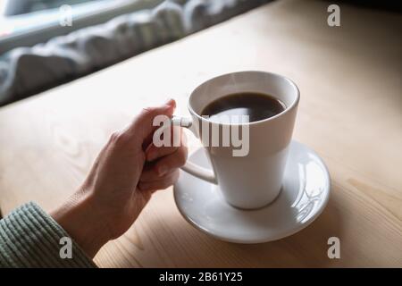 Black coffee in a white mug and saucer being held by a woman. The cuff of a cord jacket can be seen. the Table is wooden and the scene is lit with nat Stock Photo