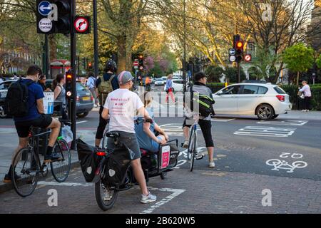 London, England, UK - April 19, 2018: Cyclists, including a Pedal Me taxi and cargo hire bike, wait at traffic lights on the Quietway 2 cycleway in Is Stock Photo