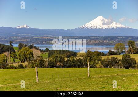 The snow-capped mountain of Volcan Osorno rises behind Lago Rupanco lake and fields of pasture in the Los Lagos region of Chile. Stock Photo
