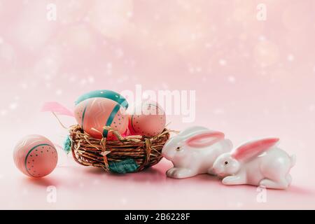 Colored decorated eggs in small basket and porcelain bunnies on pink background. Easter concept, card with free copy space. Stock Photo