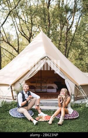 Summer picnic at sunset. Young couple wearing stylish boho casual clothes sitting on the pillows on green grass in front of big wigwam tent outdoors Stock Photo