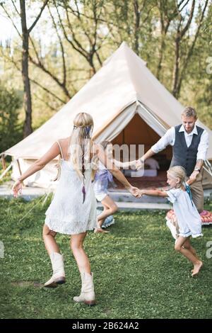 Happy active family wearing stylish boho casual clothes, dancing and jumping in green field or park in front of tipi tent. Summer vacation, family Stock Photo