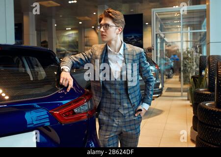 young good looking man advertising a new car, close up side view photo. pensive thoughtful businessman looking aside, leaning on blue car. Stock Photo