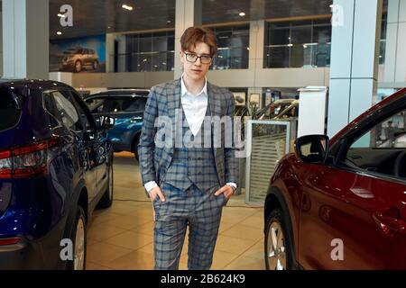 young elegant salesman standing in front of car in a dealership, guy in gray checked suit with hands on the pocket looking at the camera, man offers v Stock Photo