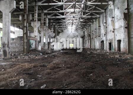 View inside an abandoned dilapidated industrial building with large space and high ceiling, with columns and light through the holes in the ceiling. The floor is covered with dark brown sand. Stock Photo