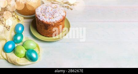 Easter festive banner with Easter cakes, colored Easter eggs on yellow cloth, willow twigs, bokeh on painted wooden background. Holiday background. Co Stock Photo