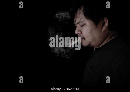 The fat man was wearing a black shirt smoking at the black background, Image of cigarette smoke spread in hand Stock Photo