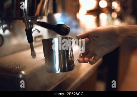 professional barista is holding a cup of milk, standing near a coffee machine, close up side view photo. Stock Photo
