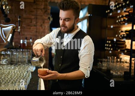 young barista in fashion vest, shirt and pants enjoying making cuppuchino at workplace, close up side view photo Stock Photo