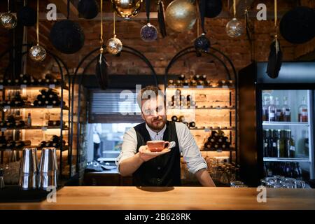 Close up of handsome bearded barista holding cup of coffee in hand looking at the camera at the cafe, close up photo.job, profession, occupation Stock Photo