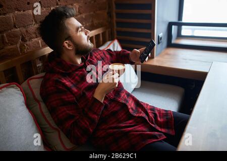 young thoughtful pensive man looking at the window, holding a smart phone and cup of tasty tea, thinking about business, life, woman Stock Photo