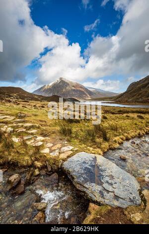 Stepping stones across melt water from snow covered Glyder Fawr leading to Llyn Idwal with Pen yr Ole Wen in the background, Snowdonia, North Wales Stock Photo