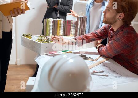 young ginger handsome man in stylish red shirt pointing to the model with a pen, talking about features of project to his colleagues during the meetin Stock Photo