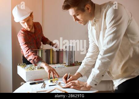 young bearded guy in helment pointing to the blueprint, giving advise to his colleague who is drawing a house on the paper in the office with modern i Stock Photo