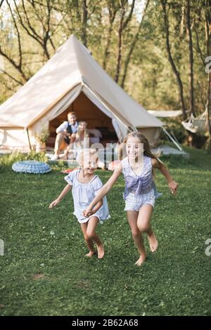 Beautiful young family spending time together in summer park or meadow, having a picnic. Young parents sitting near wigwam, while two little girls Stock Photo