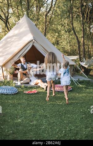 Happy family with two female children outdoors near the tent wigwam tipi in boho rustic style. Handsome man and pretty woman parents sitting near tent Stock Photo