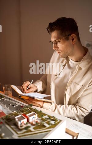 cheerful handsome architect is writing his ideas on the notebook, designer enjoying his work. close up side view photo. plan, guy found inspiration , Stock Photo