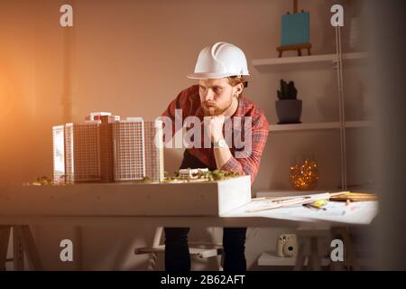 young professional pensive thoughtful architect leaning on his arm, thinking about the project, looking at the breadboad model in the studio. close up Stock Photo