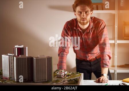 confident serious bearded architect with a breadboard model of a building looking at the camera, leaning on the table, posing to the camera Stock Photo