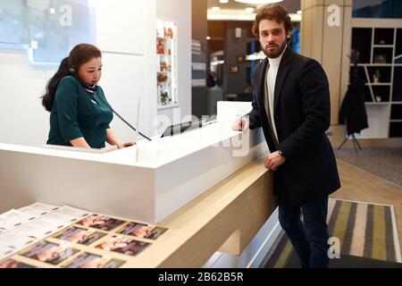 bearded handsome man looking at the camer while leaning on the reception's desk. woman talking on the phone, guy odering breakfast, dinner, lunch Stock Photo