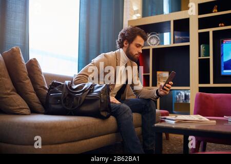 handsome bearded business man with suitcase sitting in waiting room, close up side view photo. device, gadget concept, serious man concentrated on wor Stock Photo