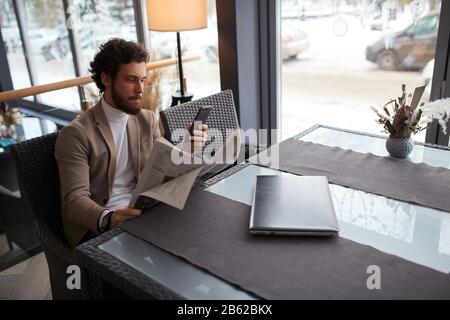 young serious man in fashion beige jacket and roll neck sitting at the table, making a phone call, chatting online with clients, dowload music, video, Stock Photo