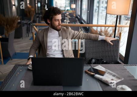 relaxed man sitting at the cafe table with a cup of coffee and laptop, looking at the window, waiting for partners, close up photo Stock Photo