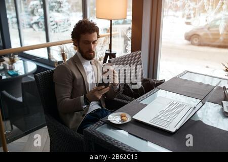 bearded pleasant man is concentrated on texting sms, best friend, man holding a cup and using smart phone . close up photo Stock Photo