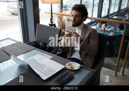 young bearded male having pensive expression, sitting at the table and thinking about success Stock Photo