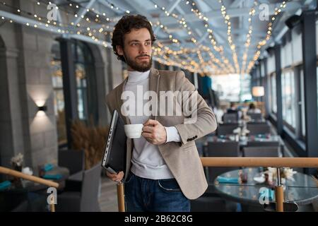 pleased cheerful young man wearing fashion jacket, jeans, roll neck holding cup of coffee and shut laptop in city cafe, looking aside, away, success, Stock Photo