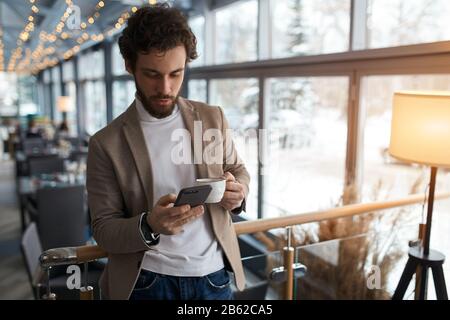 young man sending message to colleague. confident attractive young man holding smart phone and looking at it while standing in front of the big window Stock Photo