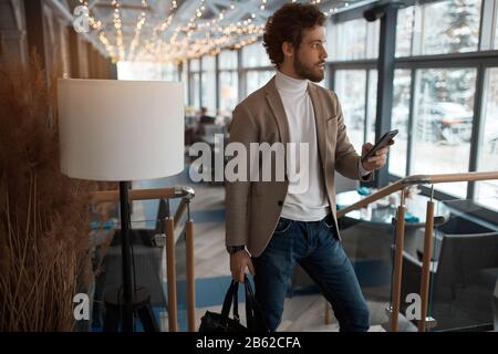 Urban business man using smart phone traveling walking inside the airport. Casual young businessman wearing suit jacket looking aside holding leather Stock Photo