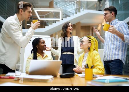 young diverse colleagues having coffe break, men drinking tea, holding cups of cofee and talking with attractive women in the office room Stock Photo
