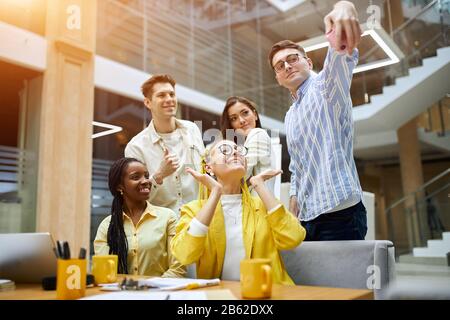 successful young funny people posing to the camera, and some guy in glasses holding gadget and asking his friends to look at the screen. modern office Stock Photo